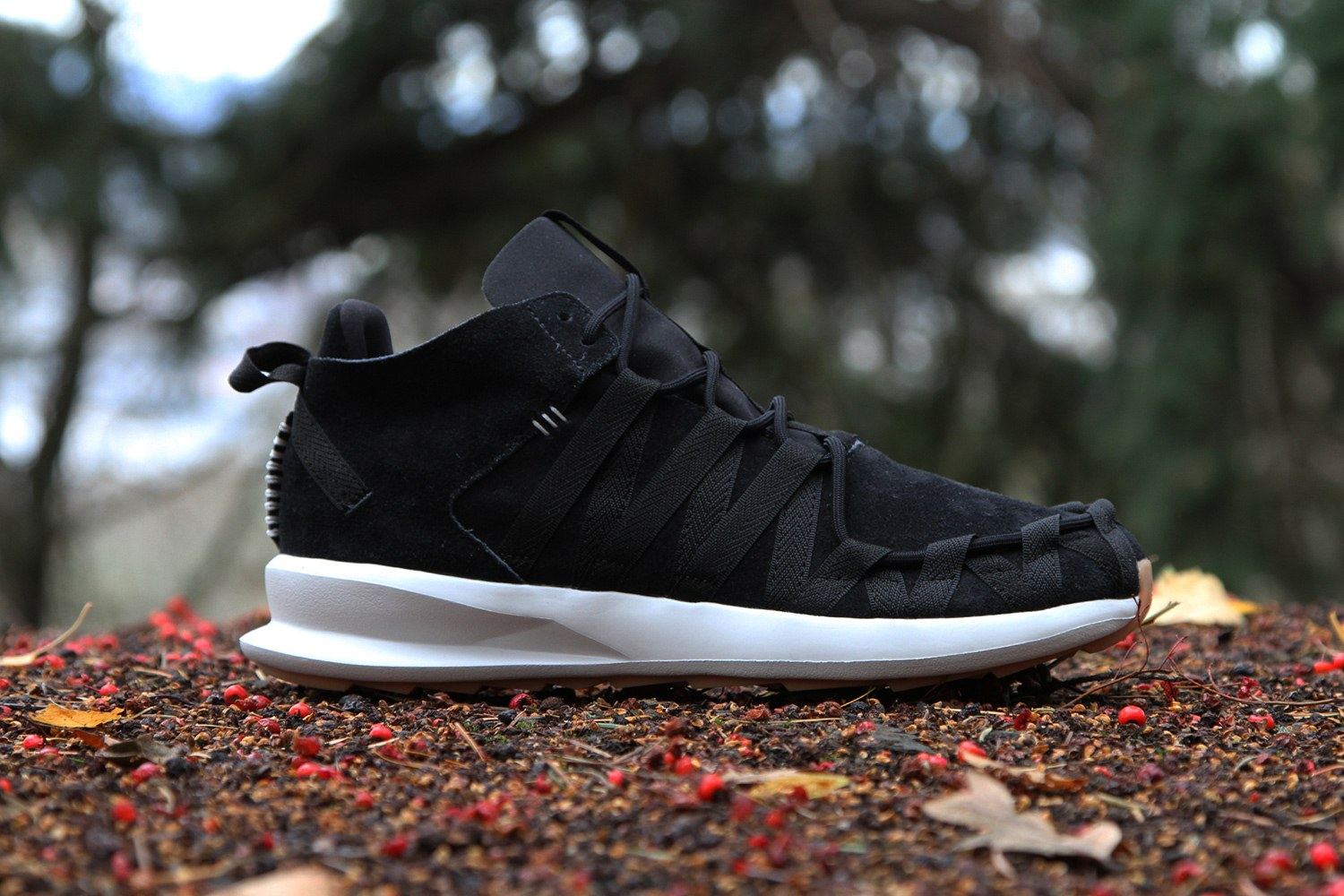 The black SL Loop Moc from the side on a bed of leaves, in black suede with black webbing straps, a white midsole and a gum heel.
