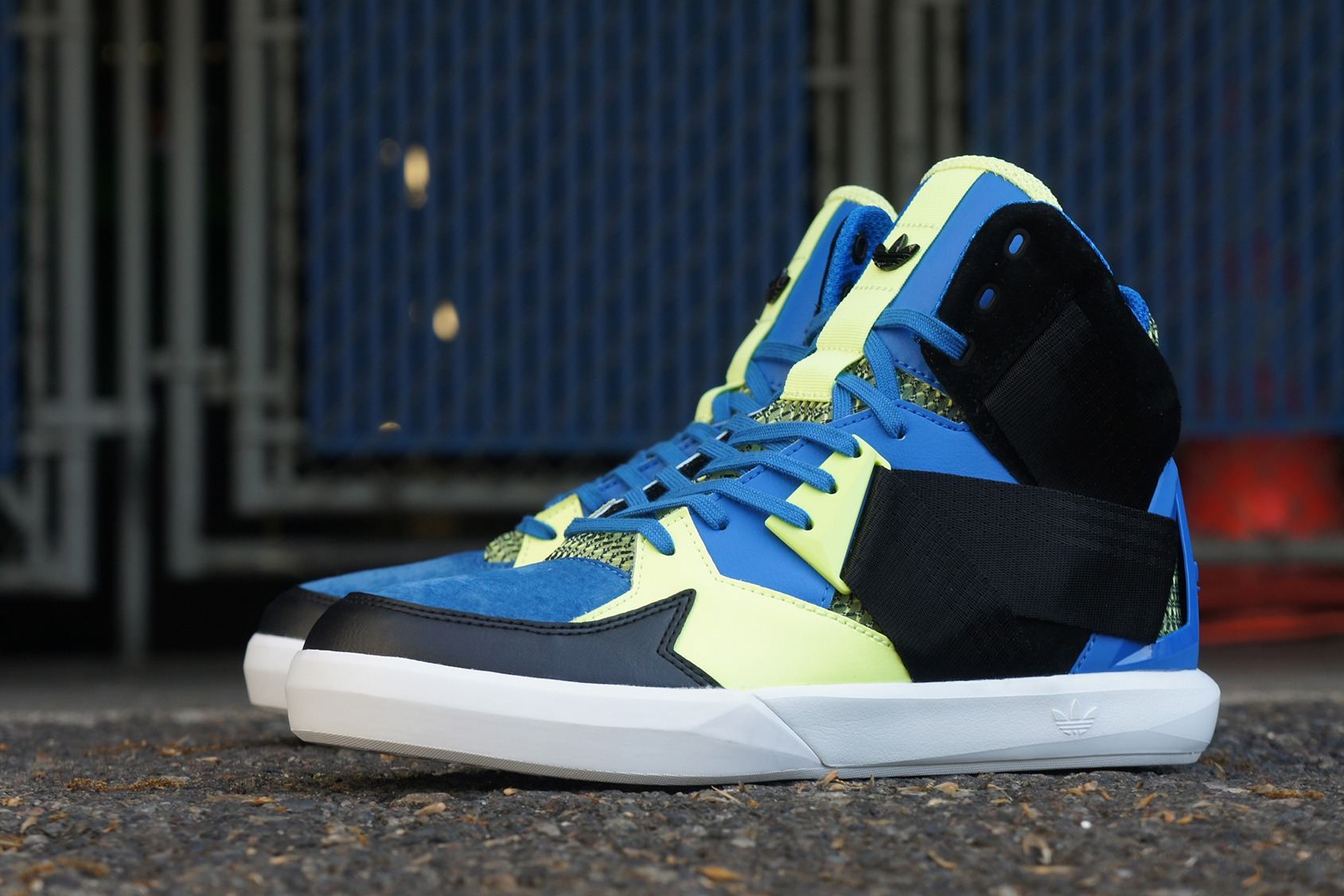 The C-10 in blue and volt with a black strap and white midsole, on pavement in front of a blue fence.