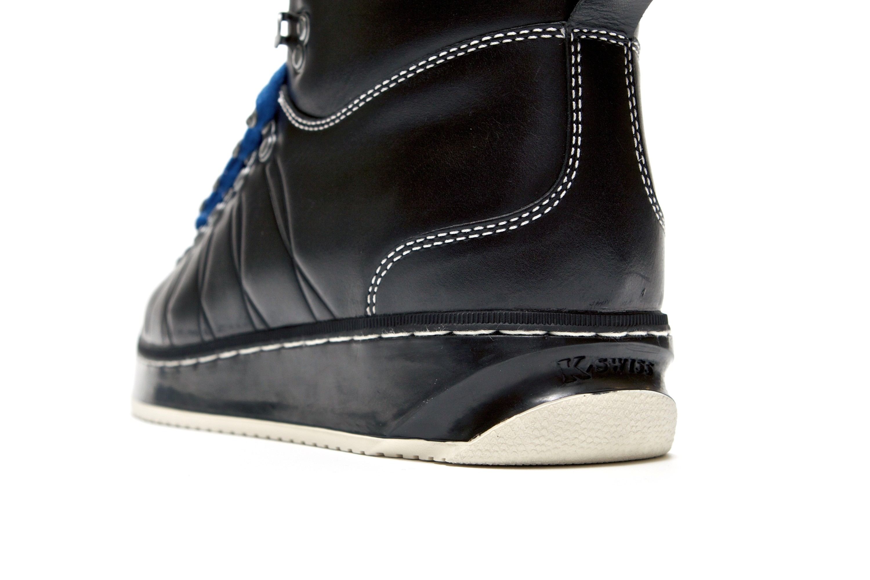 Close-up of the black boot's heel, white contrast stitching over smooth leather, the molded K-Swiss logo on the black sidewall and a cream lug sole.