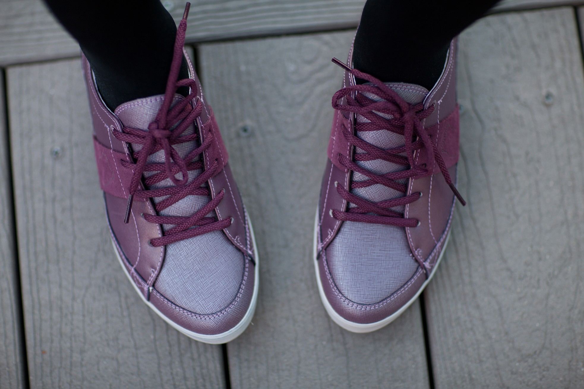 Looking down at a pair of eggplant and lilac City Tennis sneakers worn on a grey wooden deck.