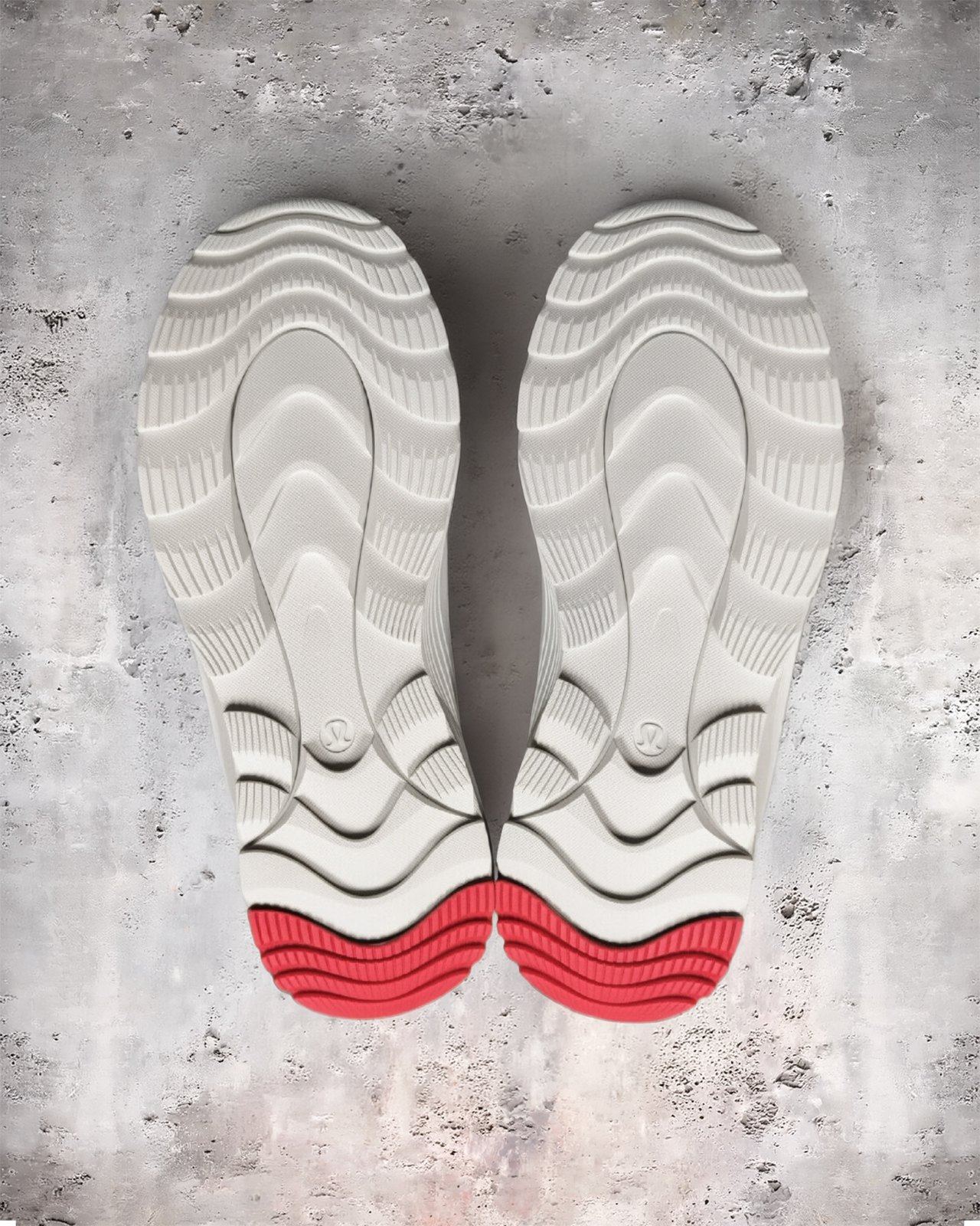 Outsoles of a pair of cream boots with wave-shaped lugs and red heel rubber, seen from above on concrete.