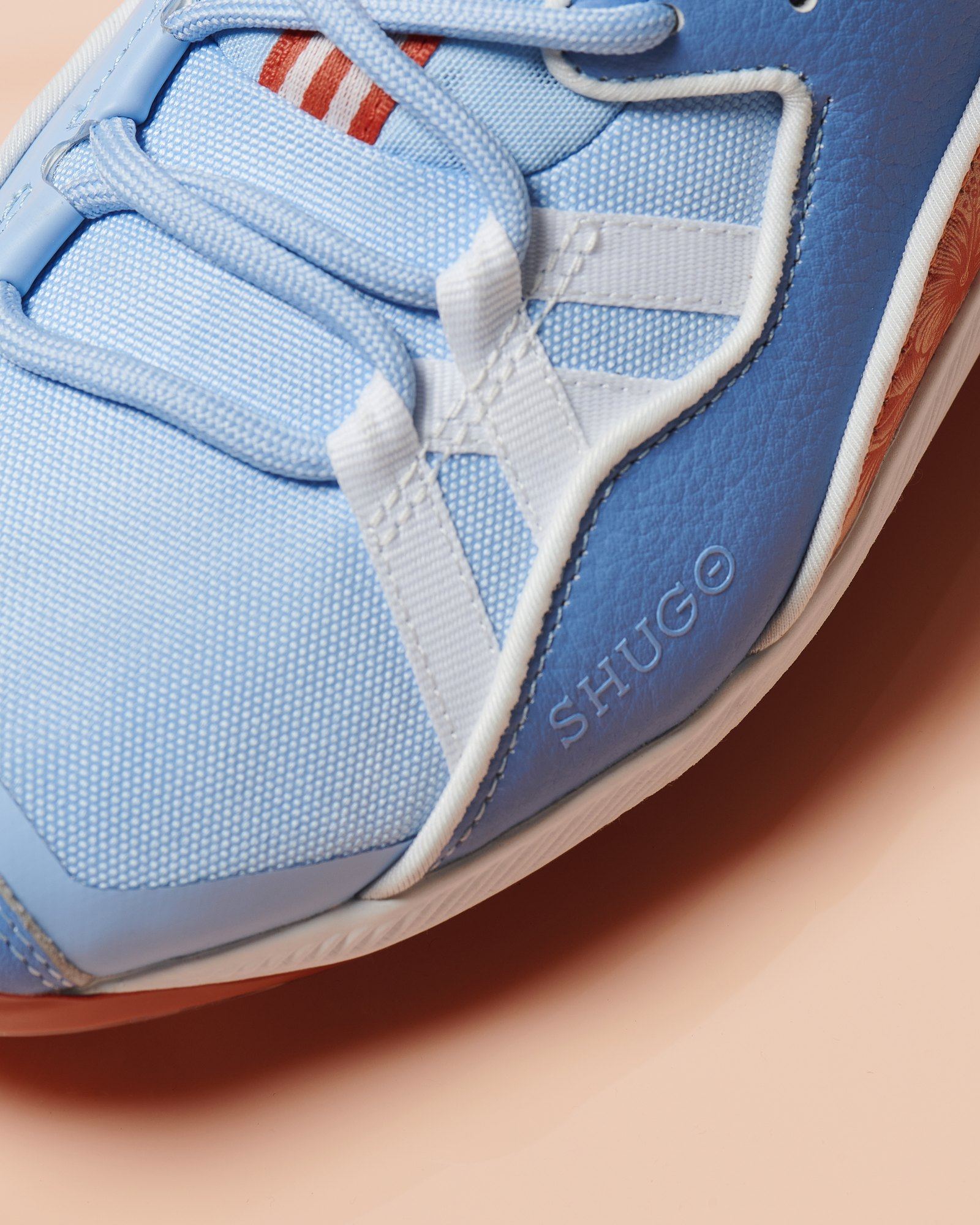 Close-up of the sky blue forefoot with white lace cage and SHUGO wordmark, on peach.