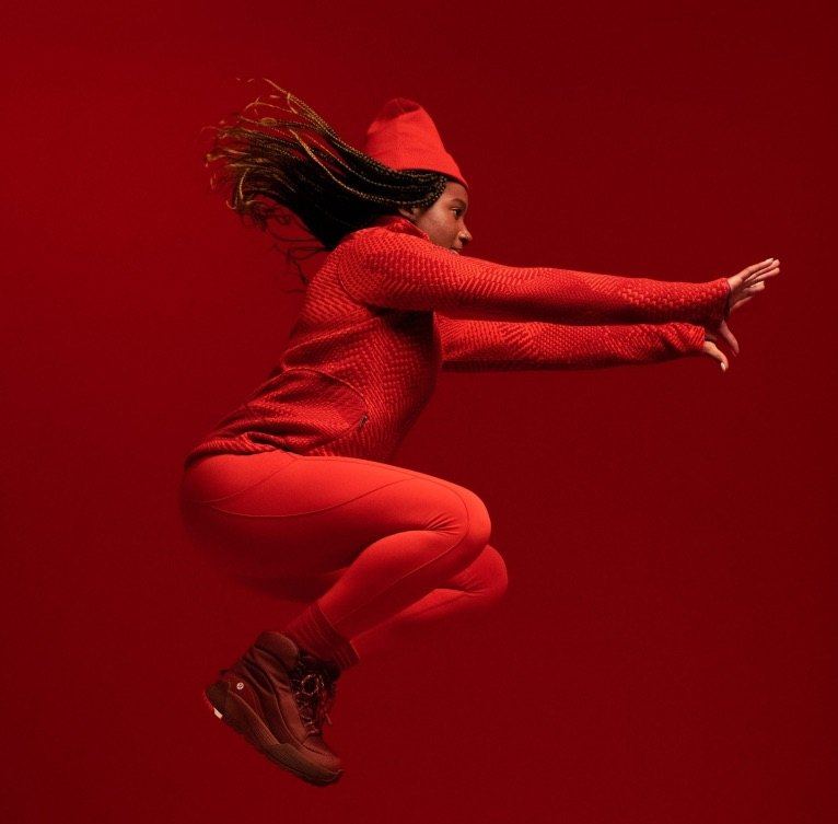 An athlete in a red knit top, leggings, and beanie mid-jump against a red backdrop, wearing the red boots.