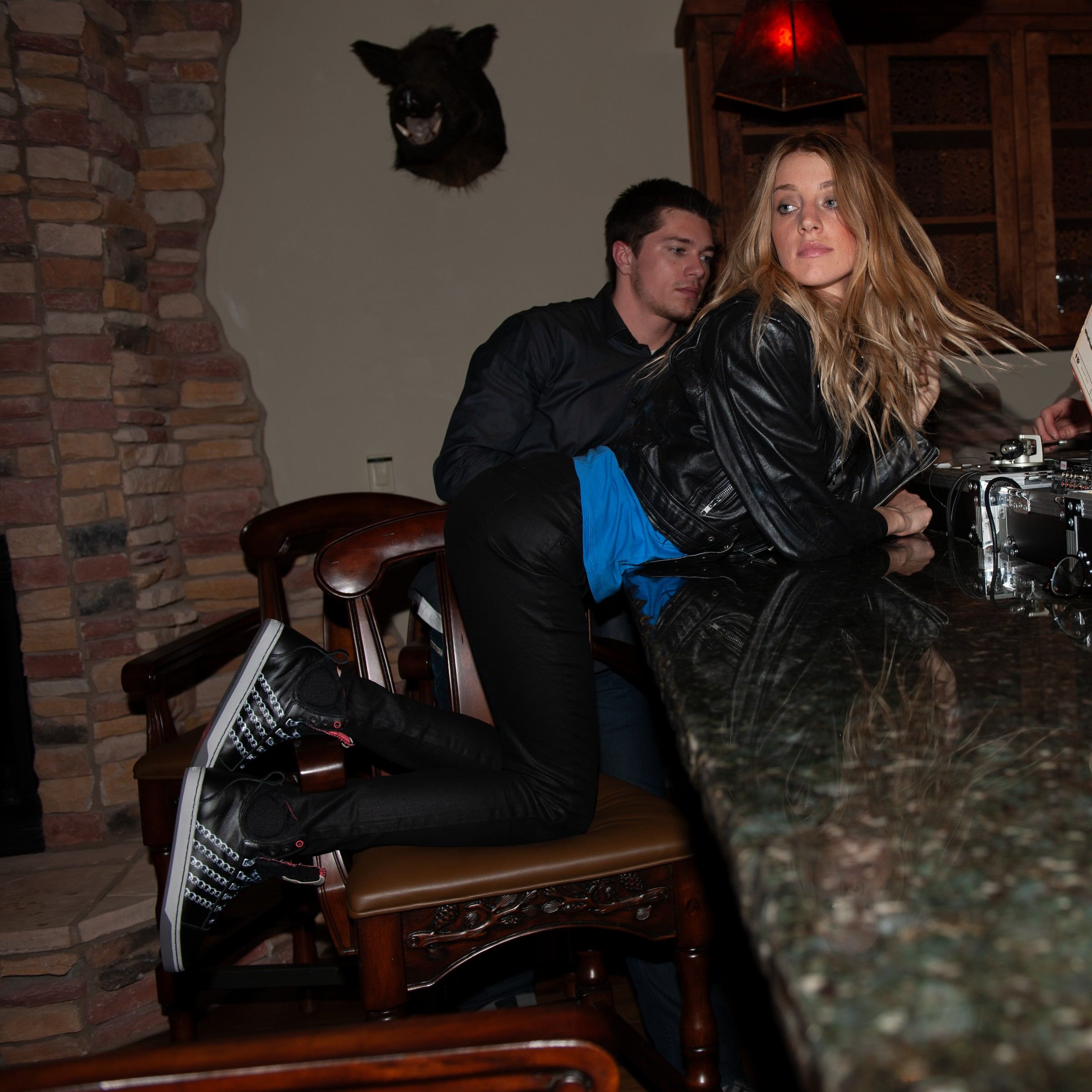 A woman in a leather jacket kneeling on a bar stool in a lodge, wearing black Slyon mids with silver chain stripes, a man seated behind her.