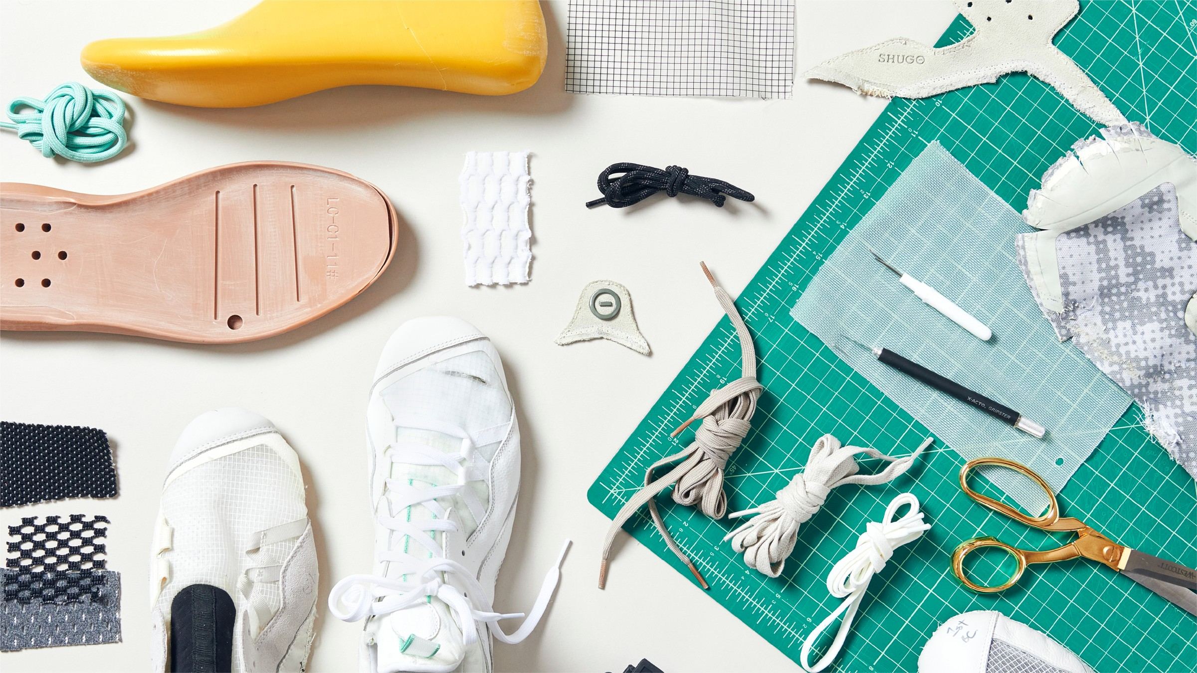 Flat lay of cleat materials and tools on a green cutting mat, with a yellow last, a pink sole pattern, white sample uppers, mesh swatches, laces, a rose gold logo badge, and scissors.