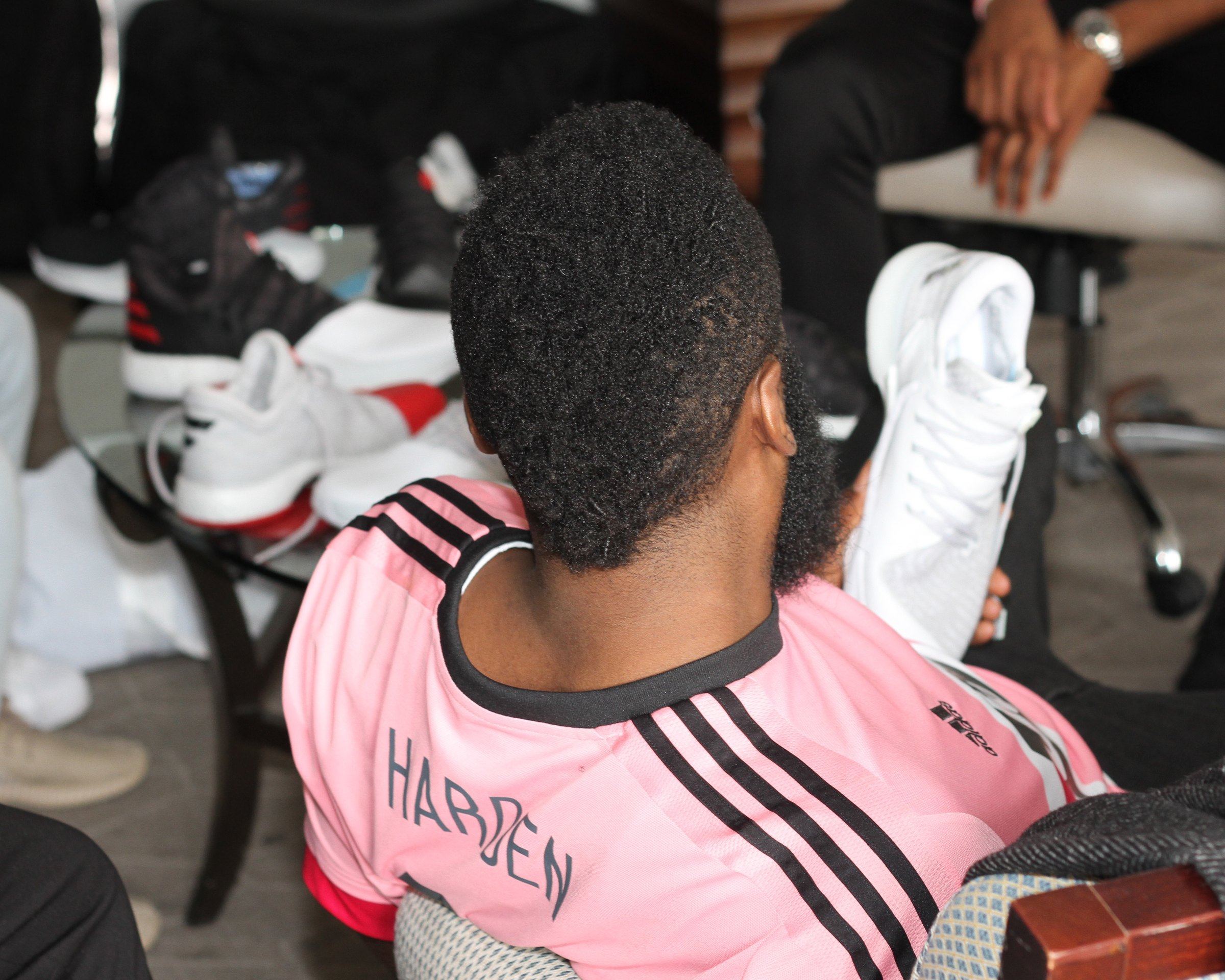 James Harden, seen from behind in a pink jersey, holding a white sample while other sample pairs sit on the table in front of him.